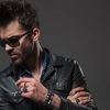 young hipster wearing rings, sunglasses and leather jacket looks down at something on grey background
